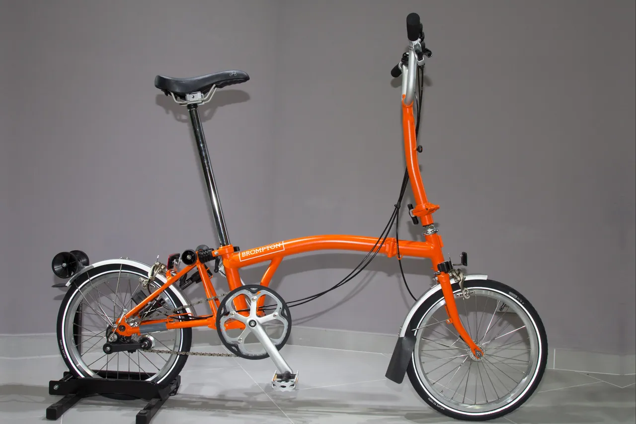 Orange Brompton folding bicycle, unfolded, with small wheels and mudguards, standing in a rear-wheel stand