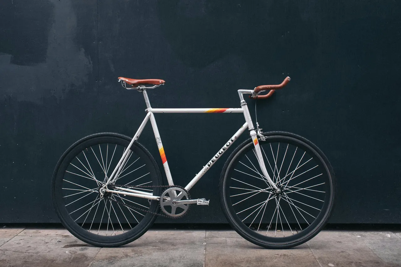 White fixed-gear bike with a single chainring and a brown leather saddle against a dark wall